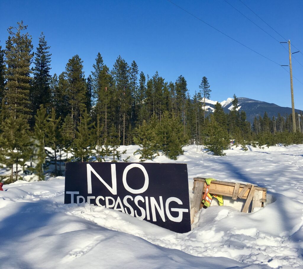A sign in the foreground reads "no trespassing." The ground is covered in snow, with conifer trees and snow-covered mountains in the distance. A transmission line can also be seen above the trees.
